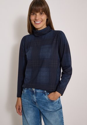 Femme souriante aux cheveux bruns portant un col roulé bleu foncé à motifs et un jean bleu, une main dans la poche, sur fond uni.