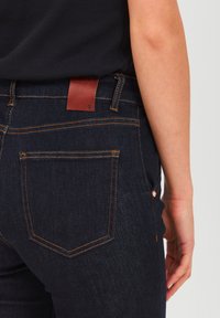 Dark blue denim jeans with orange stitching, featuring back pockets and a brown leather tag at the waistband. Fitted design, smooth texture.