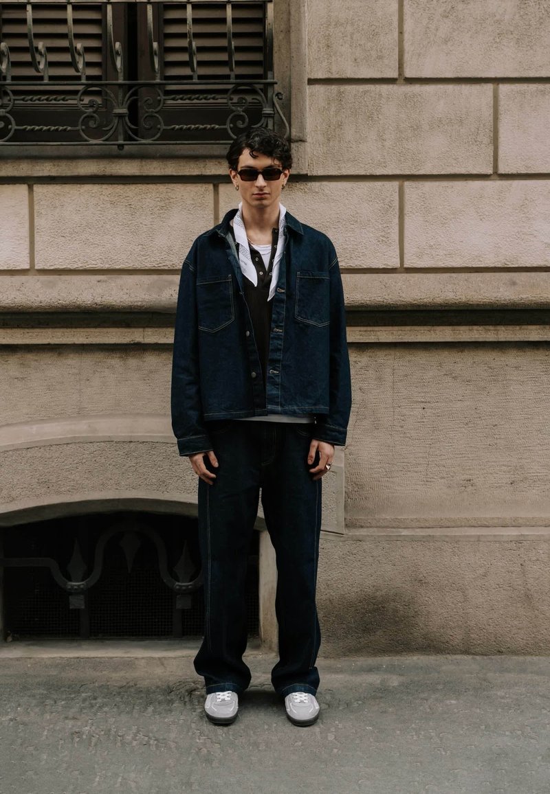 Jeune homme portant une veste et un pantalon en jean foncé, des baskets blanches, des lunettes de soleil et une écharpe, debout contre un mur en pierre beige avec une fenêtre.