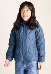 Young girl with long dark hair wearing a blue quilted jacket and blue jeans, smiling against a plain light background.