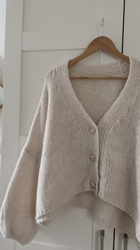 Hellbeige gestrickter Cardigan mit drei Knöpfen, der an einem Holzbügel vor einer weißen, vertäfelten Tür hängt.