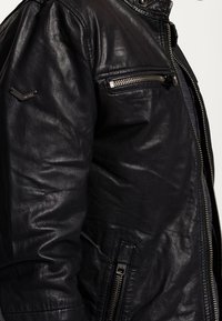 Giacca in pelle nera con una texture liscia, dotata di tasca frontale con zip, accessori metallici e colletto alto per un ulteriore calore.