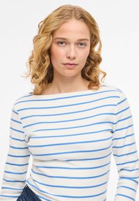 Νεαρή γυναίκα με σγουρά ξανθά μαλλιά, ντυμένη με λευκό πουκάμισο μακρυμάνικο με ανοιχτό μπλε οριζόντιες ρίγες, στέκεται μπροστά σε ένα απλό φόντο.