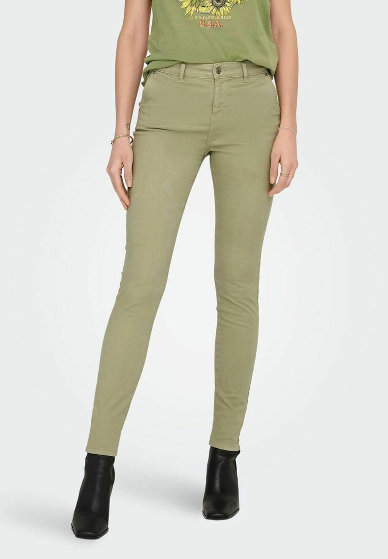 Femme portant un pantalon slim vert olive, un t-shirt vert graphique et des bottines noires, debout sur un fond clair.