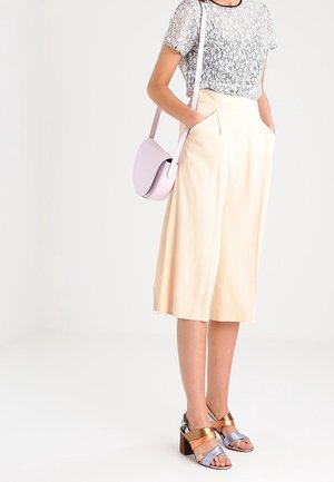 Lichtbeige culotte-broeken gecombineerd met een transparante, gestreepte grijze top. Het model draagt een lichtroze schoudertas en heeft gekleurde haksandalen aan.