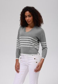 Pulover gri tricotat cu guler în V, având dungi orizontale albe. Asortat cu pantaloni albi, care prezintă accente cu nasturi lângă buzunare.