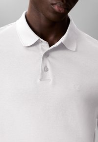 White polo shirt made of soft fabric, featuring a classic collar, three buttons, and an embroidered logo on the left side.