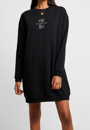 Nainen pukeutunut mustaan Calvin Klein -collegepukuun, jossa hopeinen "cK Calvin Klein 50" -logo, pitkät hihat ja pyöreä pääntie.