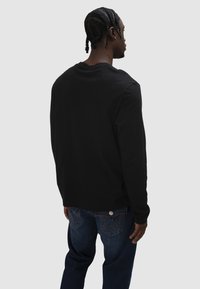 Mann mit geflochtenen Haaren trägt ein schwarzes Langarmshirt und dunkelblaue Jeans, steht mit dem Rücken zum Betrachter vor einem schlichten hellen Hintergrund.