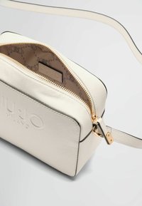 Sac bandoulière en faux cuir blanc avec fermeture éclair dorée, logo en relief et intérieur à motifs. Accentué par une fine bandoulière et une forme rectangulaire.