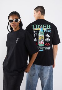 T-shirts noirs surdimensionnés, l'un avec un imprimé graphique d'une bouteille de soda présentant un tigre, et l'autre avec un petit logo sur la poitrine.