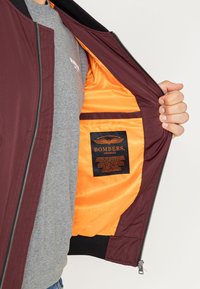 Persoon houdt een open bordeauxrode bomberjas vast met een feloranje voering en zichtbaar label "Bombers Original" aan de binnenkant.