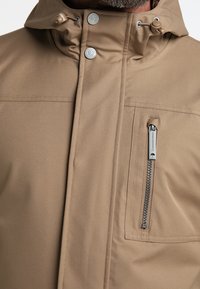 Vedenkestävä beige takki, jossa on korkea kaulus, napit, vetoketjullinen rintatasku ja sileä rakenne, johon on ommeltu yksityiskohtia.