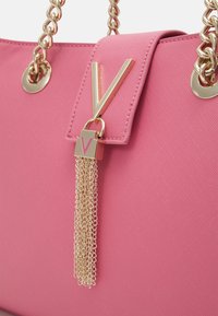 Rosa handväska med texturerad yta, som har ett guldhänkel, en stor V-formad accent och en gyllene kvastdetalj.