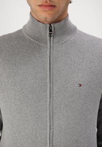 Sivá mikina na zips s vysokým golierom, vyrobená z textúrovanej pleteniny. Obsahuje malé logo na ľavej hrudi a hladký zips.