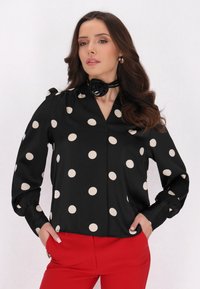 Blusa nera con grandi pois crema, con scollatura a V e maniche lunghe, abbinata a pantaloni rossi. Include un dettaglio di choker floreale nero.