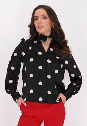 Blusa nera con grandi pois crema, con scollatura a V e maniche lunghe, abbinata a pantaloni rossi. Include un dettaglio di choker floreale nero.