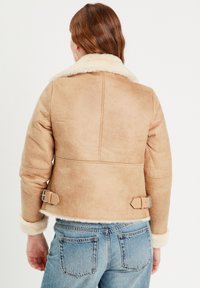 Chaqueta de piel sintética de color marrón claro con un cuello crema, diseño recortado y detalles de hebillas en los lados. Textura suave, mangas de corte recto.
