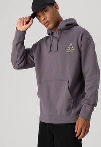 Sudadera con capucha gris, bolsillo frontal, capucha ajustable con cordón y un logo en forma de triángulo impreso en color crema. Tela suave y corte relajado.