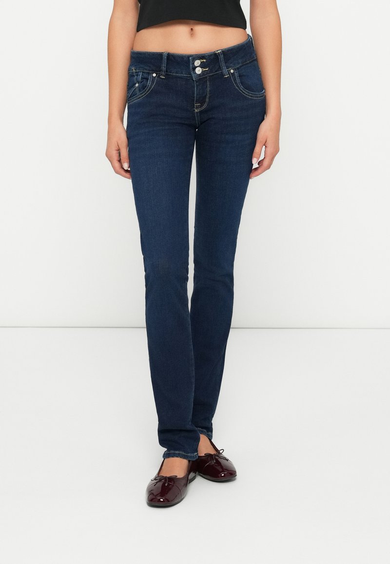 Mujer vestida con jeans ajustados de color azul oscuro y bailarina de charol burdeos, de pie contra un fondo blanco liso.