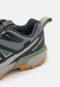 Szare i zielone buty turystyczne z teksturowaną cholewką, gumową podeszwą i uchwytem. Posiadają logo GORE-TEX oraz wyraźny wzór bieżnika.