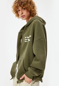 Olijfgroene ritsshirt met een losse pasvorm, grote capuchon, voorzakken en witte tekst die zegt "do WHERE you WILD all THE."