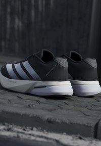 Paire de chaussures de course Adidas noires et blanches avec trois bandes, posées sur une surface pavée en extérieur.