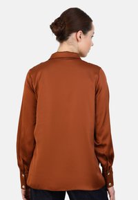 Braune Bluse mit langen Ärmeln aus glattem, glänzendem Stoff, mit Kragen und geknöpften Manschetten. Leicht geschwungener Saum hinten.