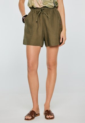 Kobieta ubrana w oliwkowe szorty ze ściągaczem, wzorzysty top, brązowe sandały wsuwane oraz ciemną bransoletkę na lewej ręce.