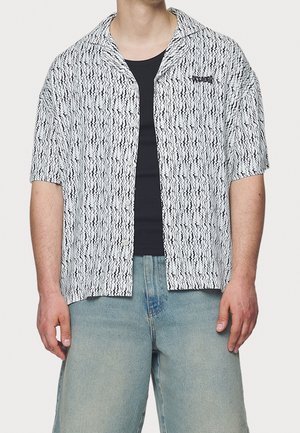 Camisa de manga corta con botones en un patrón ondulado en blanco y negro, con collar y logotipo en el pecho izquierdo, llevada sobre una camiseta de tirantes negra. Jeans de color azul claro.