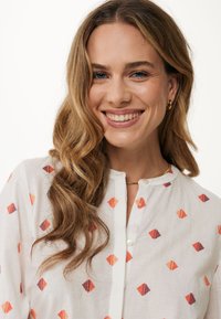 Camicia bianca con motivi geometrici arancioni e viola, realizzata in tessuto leggero. Presenta una patta con bottoni e un collo rotondo.