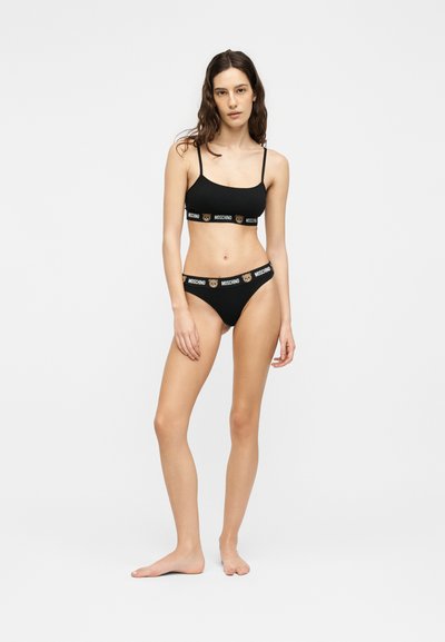 Νεαρή γυναίκα που στέκεται ξυπόλητη, φοράει μαύρα εσώρουχα Moschino με λογότυπα αρκουδάκια στις ταινίες, πάνω σε απλό λευκό φόντο.