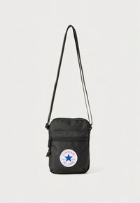 Svart nylon crossbody-veske med justerbar stropp, glidelåslomme foran og en rund Converse All Star-logo i rødt, hvitt og blått.