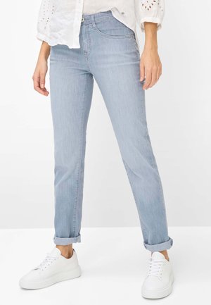 Persona che indossa jeans azzurri con risvolti, una camicetta bianca in tessuto a occhiello e scarpe da ginnastica bianche con lacci, in piedi contro uno sfondo bianco uniforme.