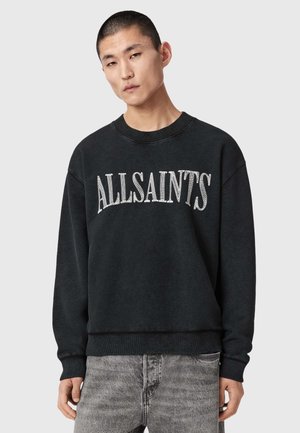 Young man wearing black sweatshirt with white "ALLSAINTS" text and faded gray jeans, standing against plain light gray background.
