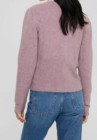 Kvinna med långt mörkt hår iförd en mauve färgad tröja och blå jeans, vänd bort med avslappnade händer vid sidorna mot en neutral bakgrund.