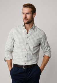 Licht geruite blouse in wit, groen en blauw; met knopen kraag; lange mouwen opgerold bij de manchetten; gecombineerd met donkere broek en een bruine riem.