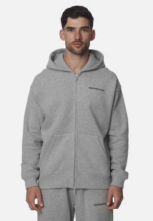 Uomo che indossa una felpa con cappuccio grigia con zip e pantaloni della tuta grigi abbinati con piccoli loghi neri con testo sul petto e sulla coscia.