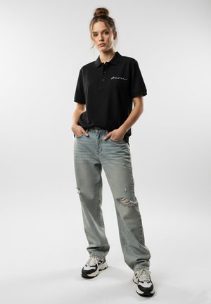 Zwarte polo met korte mouwen, met een wit geborduurd logo. Lichtblauwe versleten jeans met scheuren, gecombineerd met zwart-witte sneakers.