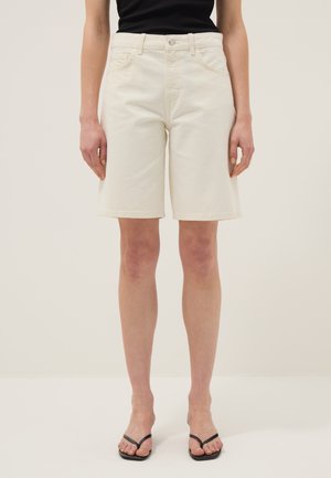 Frau trägt hoch taillierte, knielange, elfenbeinfarbene Shorts, schwarzes ärmelloses Oberteil und schwarze Flip-Flop-Sandalen und steht vor einem schlichten Hintergrund.