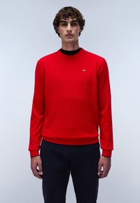 Jersey de cuello redondo rojo hecho de una suave tela de punto, con mangas largas y puños acanalados, combinado con pantalones oscuros, acento de diseño minimalista.