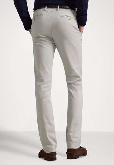 Svetlo sive chino hlače s stisnjeno prileganje, diskretnimi detajli na zadnjem žepu in gladko teksturo tkanine. Združene z rjavimi semiš čevlji.
