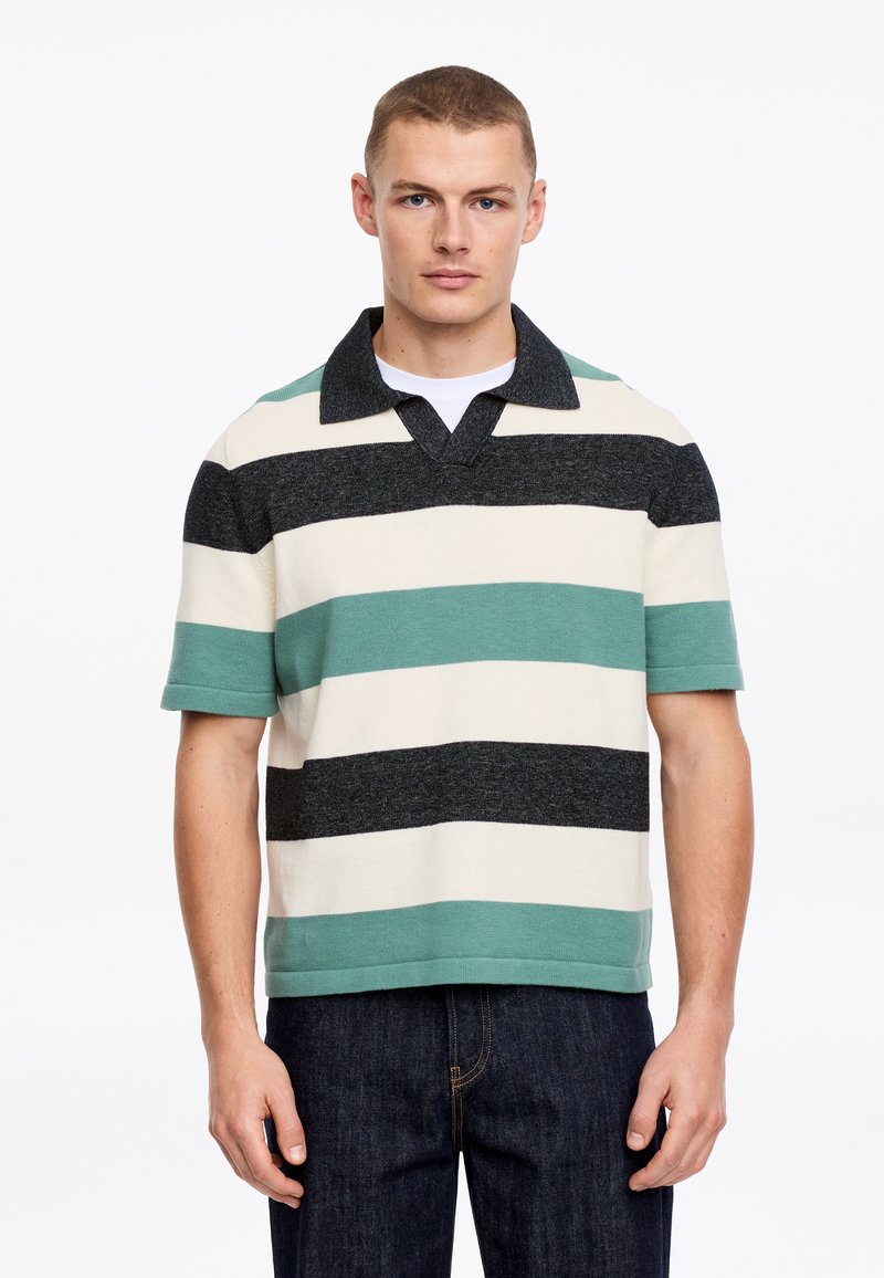 Jeune homme aux cheveux courts portant un polo à manches courtes avec de larges rayures horizontales noires, blanches et teal, et un jean foncé.