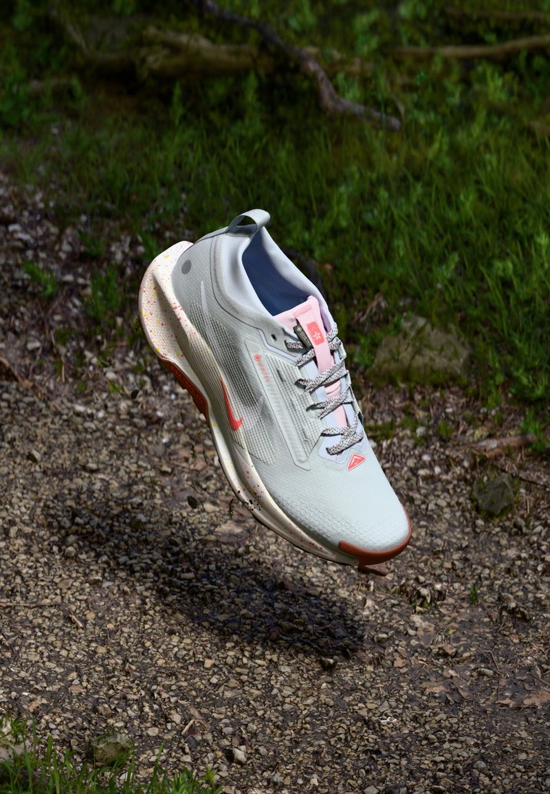 Jasnoszare sportowe buty ze różowymi akcentami, teksturowana siateczka u góry, pleciona podeszwa pośrednia i odblaskowe detale. Unoszące się nad żwirową powierzchnią.