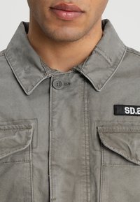 Grå bomuldsjakke med en knaplukket krave, to frontlommer og et mærke, der er mærket "SD.2." Glat tekstur og casual design.