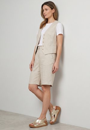 Kvinde stående op ad væg iført beige knapvest, matchende knælange shorts, hvid t-shirt og guld åbne tå-sandaler.