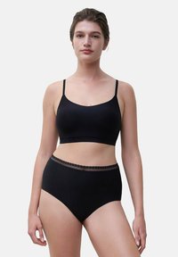 Junge Frau mit nach hinten gebundenem Haar, die ein schwarzes nahtloses Bralette und schwarze hochgeschnittene Unterwäsche mit Spitzenbesatz trägt, steht vor einem schlichten Hintergrund.