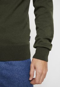 Bras droit et main d'une personne portant un pull en maille vert olive et un jean bleu, montrant l'étiquette Calvin Klein sur la couture latérale du pull.