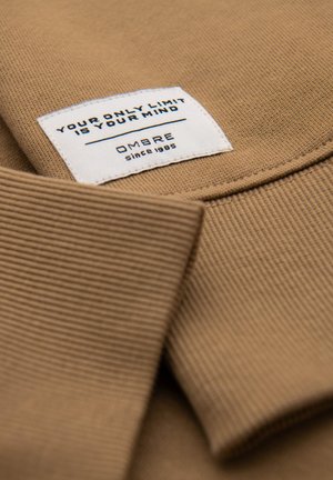 Ripustettu collegepaita, jossa on ribattua kaulusta. Valkoisessa tarrassa lukee mustalla tekstillä "AINOA RAJASI ON MIELESI" ja "OMBRE vuodesta 1985".