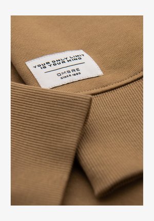 Beżowy sweter z materiału, z prążkowanym kołnierzem. Biała metka zawiera napis "TWÓJ JEDYNY LIMIT TO TWOJA GŁOWA" oraz "OMBRE od 1985" na czarnym tle.